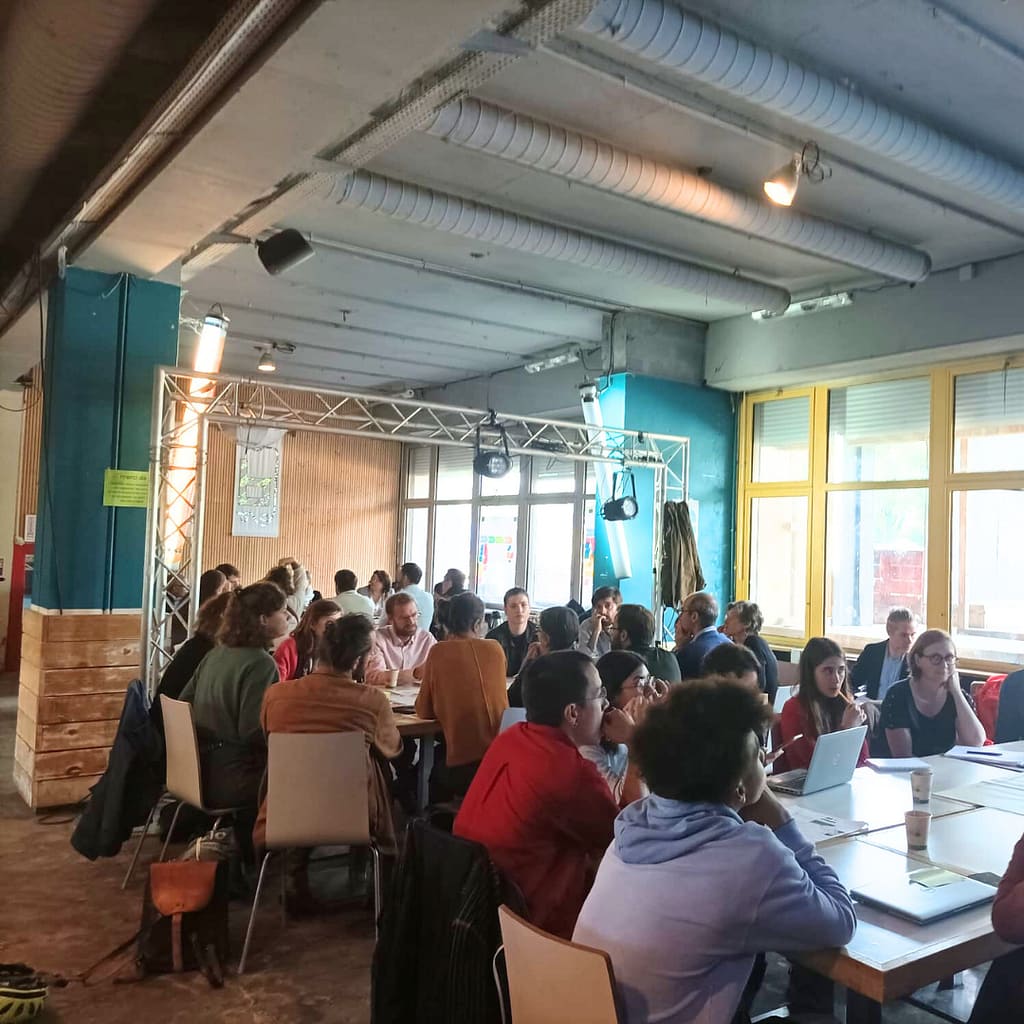 L'image montre un groupe de personnes assises autour de tables, environ 50, dans une salle industrielle