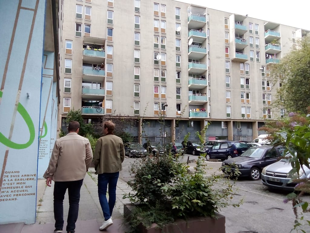 L'image montre 2 personnes de deos marchant en pied d'immeuble