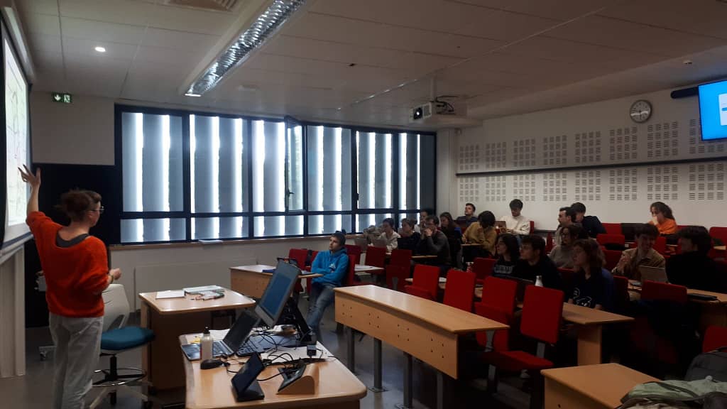 L'image montre un petit amphithéâtre dans une université, avec 4 bances de personnes assises sur des sièges rouges, et une personne debout au tableau qui présente uquelque chose