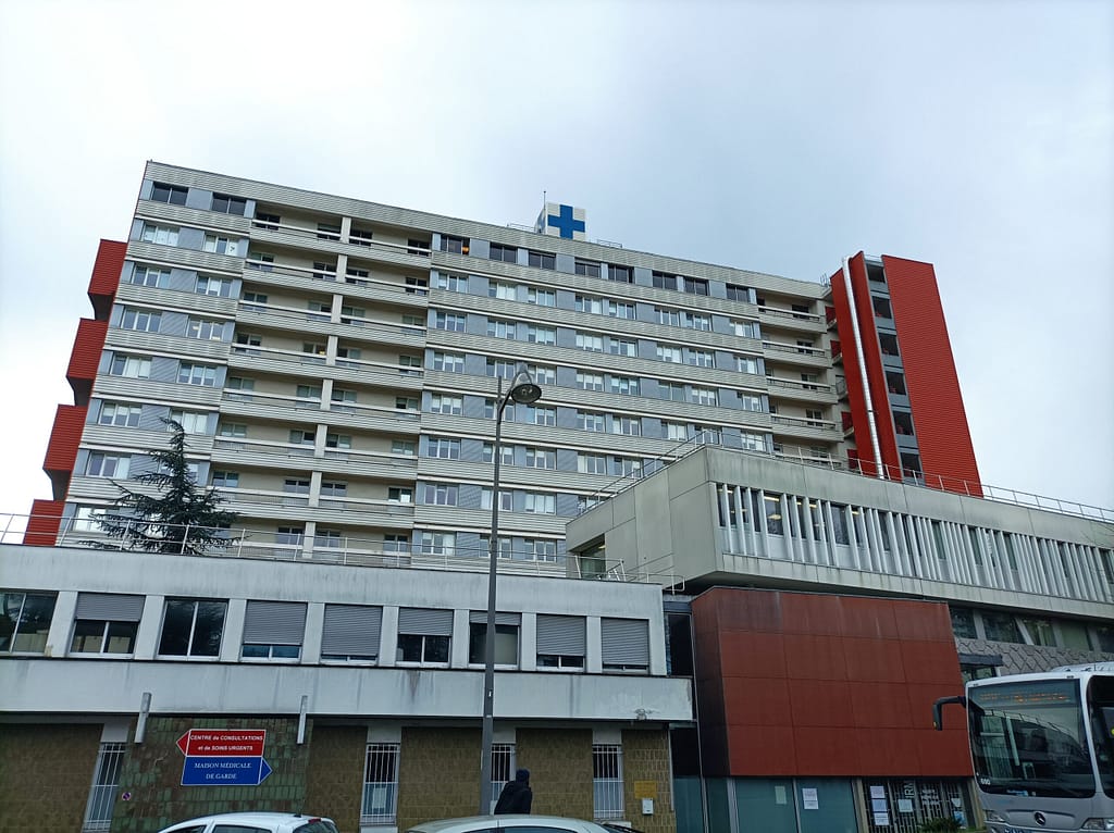 L'image montre l'hôpital de Longjumeau, de 9 étages, blanc avec des balcons, et coiffé d'une croix bleue indiquant sa fonction. 2 tours rouges l'encadrent.