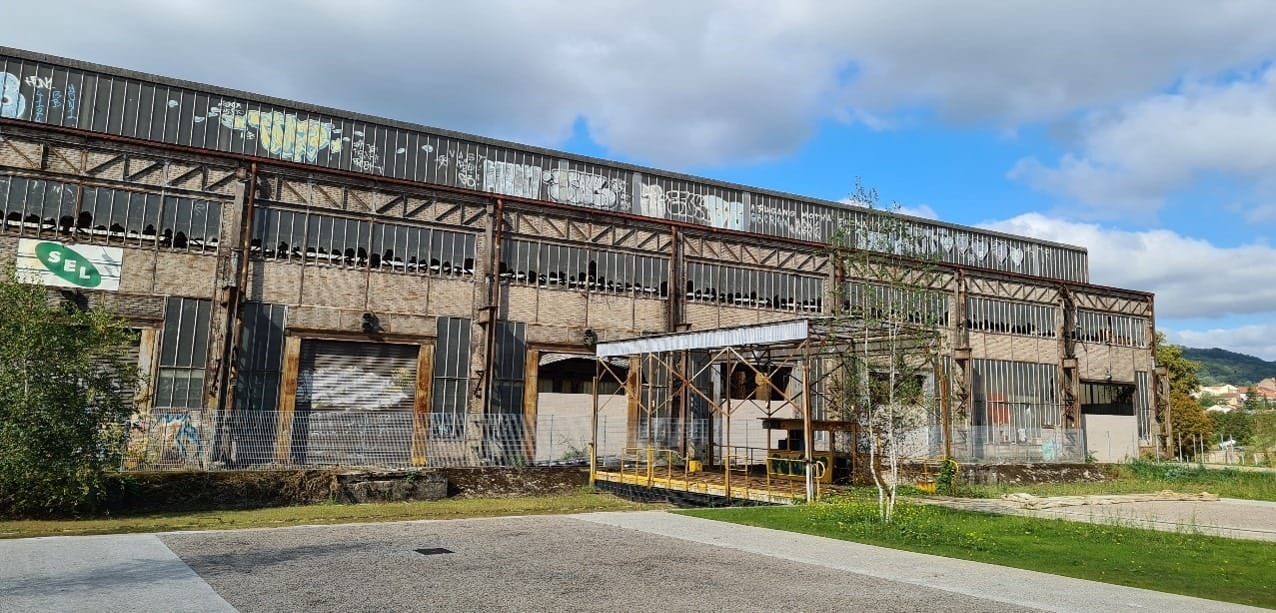 L'image montre une usine désaffectée en forme de hangar, avec des quais de déchargement pour camions recouverts de grafitis. Les ouvertures sont en mauvais état et les fenêtres cassées. Une place de parking au premier plan, du gazon, et une colline derrière