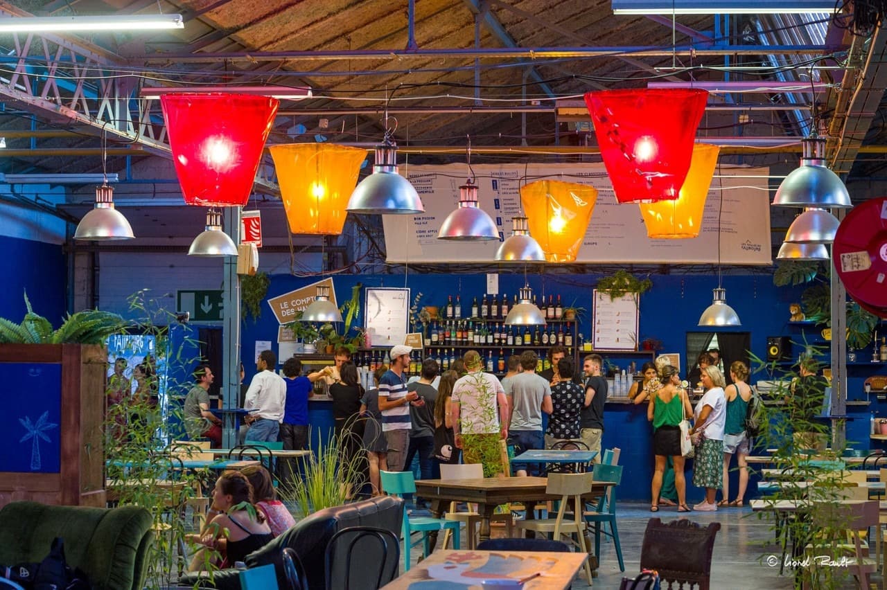L'image montre un foodcourt en intérieur, avec poutres métalliques apparentes et des grosses lampes rouges et oranges qui pendent du plafond. Des personnes sont atablées à des tables, entre des plantes. D'autres personnes sont debout au bar.
