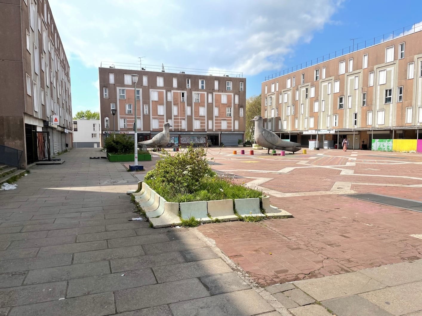 L'image montre la place aux oiseaxu de Grigny : une place au sol rouge avec des barres d'immeubles autour, et des statues de pigeons grises au centre