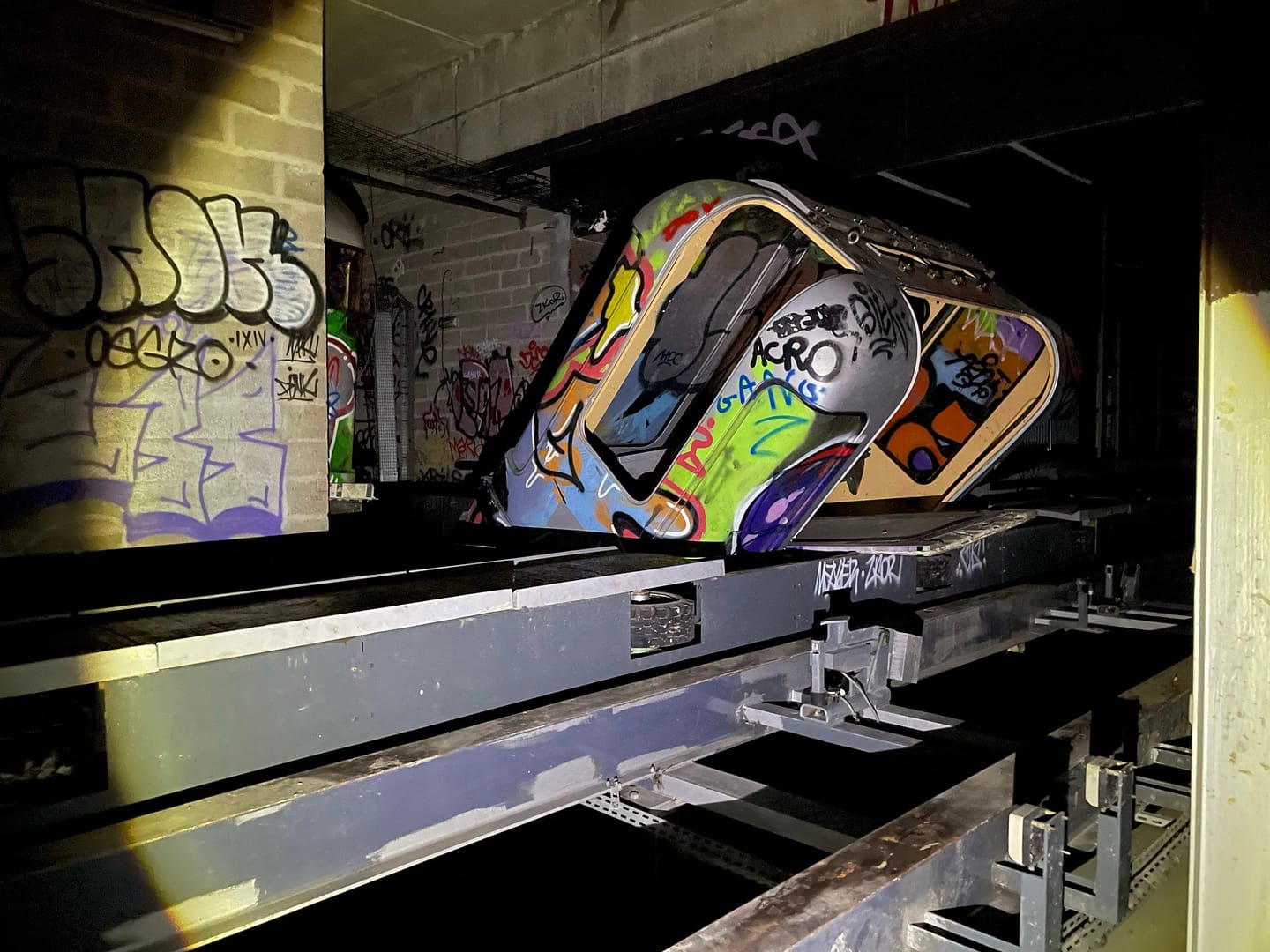 L'image montre un sous terrain tagué éclairé à la lampe, avec des rails et une rame de métro inclinée et taguée