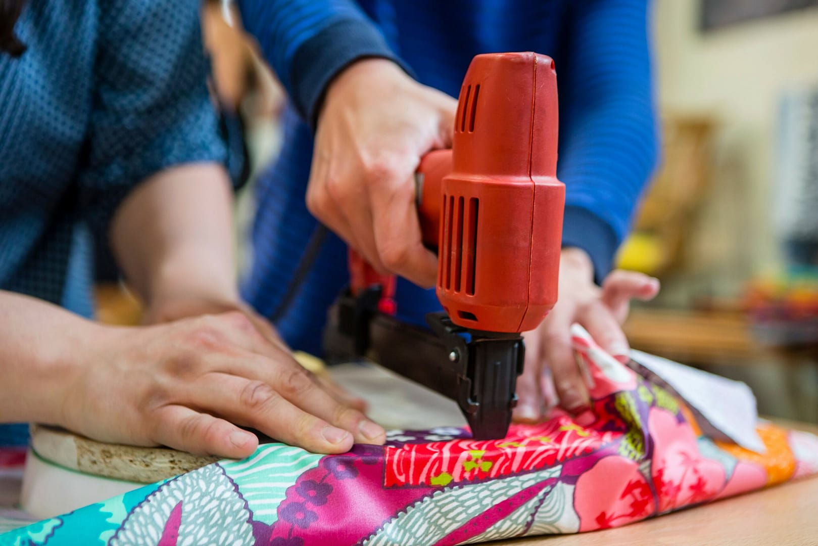 L'image montre un gros plans sur 2 mains tenant une agraffeuse à vêtements en train de coudre une veste colorée