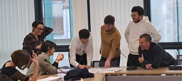 L'image montre 8 personnes assis et debout autour d'une table, ils sourient et montrent un plan