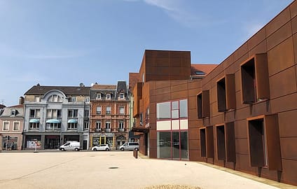 L'image montre une place publique entourée de bpatiment de maximum 2 étages, avec au premier plan un bâtiment en bois 'un étage