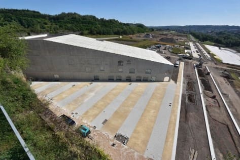 L'image montre la friche de Micheville : un zol en pente au oremier plan, un bâtiment industriel de sidérurgie, un parking derrière