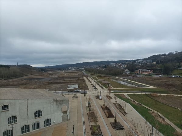 L'image montre la vue du quartier en construction depuis des hauteurs : au premier plan, un bâtiment masif gris (le centre culturel), avec un parvis bétonné blanc autour. au deuxième plan, la ZAC avec les trous creusés dans le sol pour accueillir les futurs lots