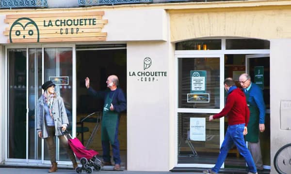 L'image montre un rez-de-chaussée avec un magasin dont le nom est inscrit en vitrine en bois : "la chouette coop". Une personne en sort avec un cadis, une autre personne avec un tablier la salue. 2 personnes marchent sur le trottoir