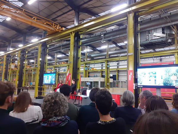 L4image montre une conférence avec des personnes assises de dos au premier plan, et 2 personnes parlant dans un micro sur la scène. Le lieu a des poutres métalliques apaprentes jaunes