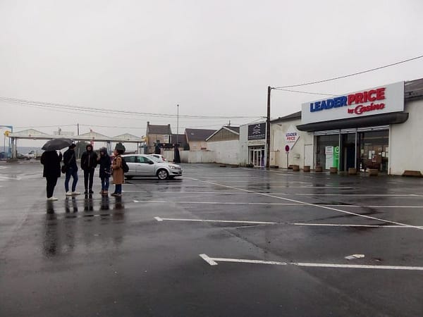 L'image montre 4 personnes sur un parking de supermarché sous la pluie