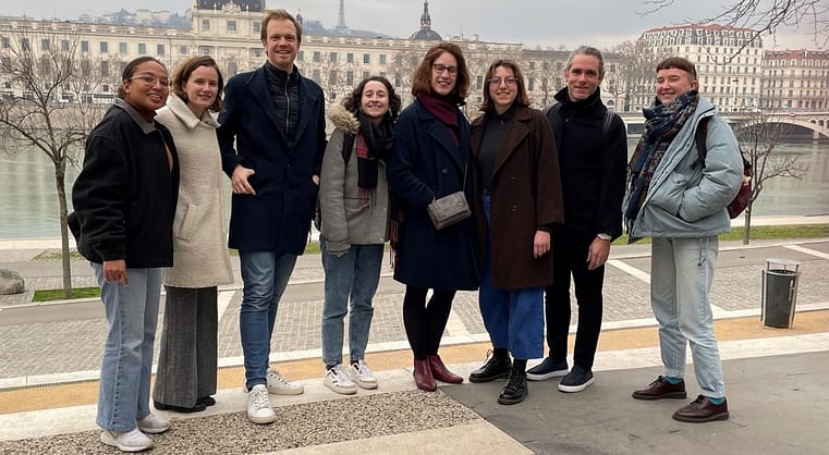 L'image montre 8 personnes debout souriant regardabt la caméra, derrière le fleuve et lyon