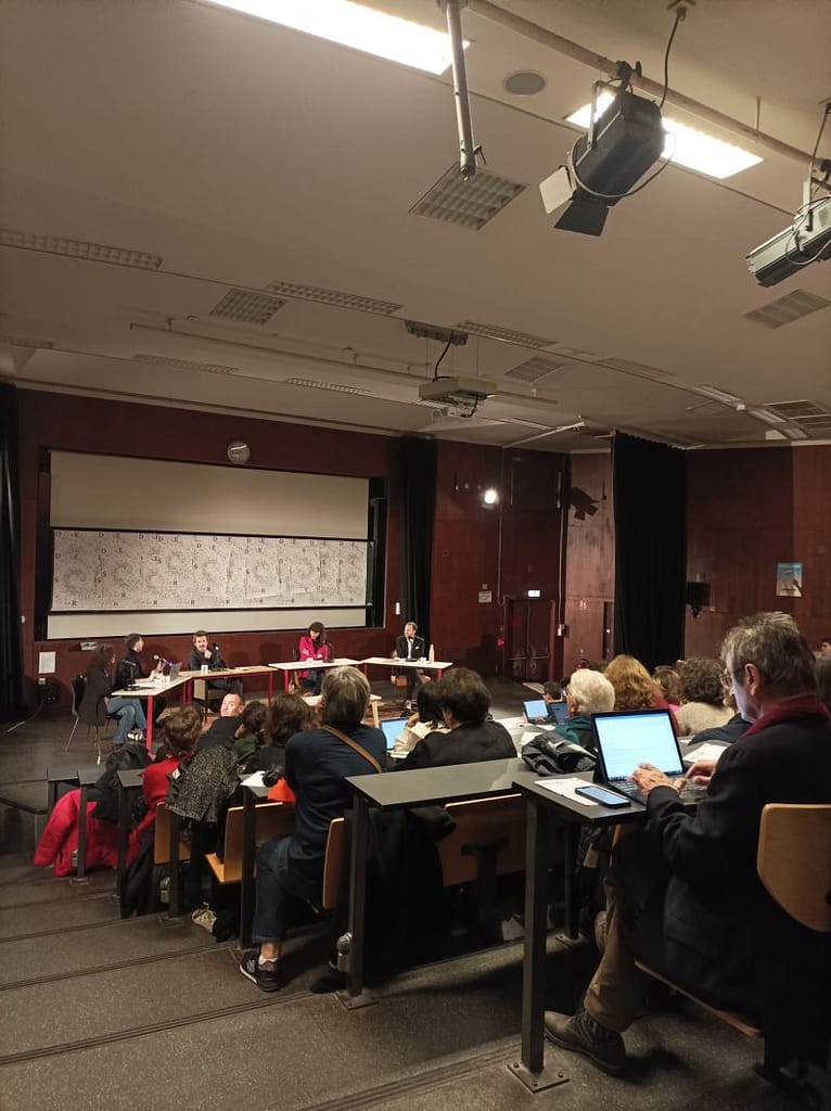 L'image montre un amphithéâtre rempli devant 4 personnes assises à des tables acvec des micro sur scène.