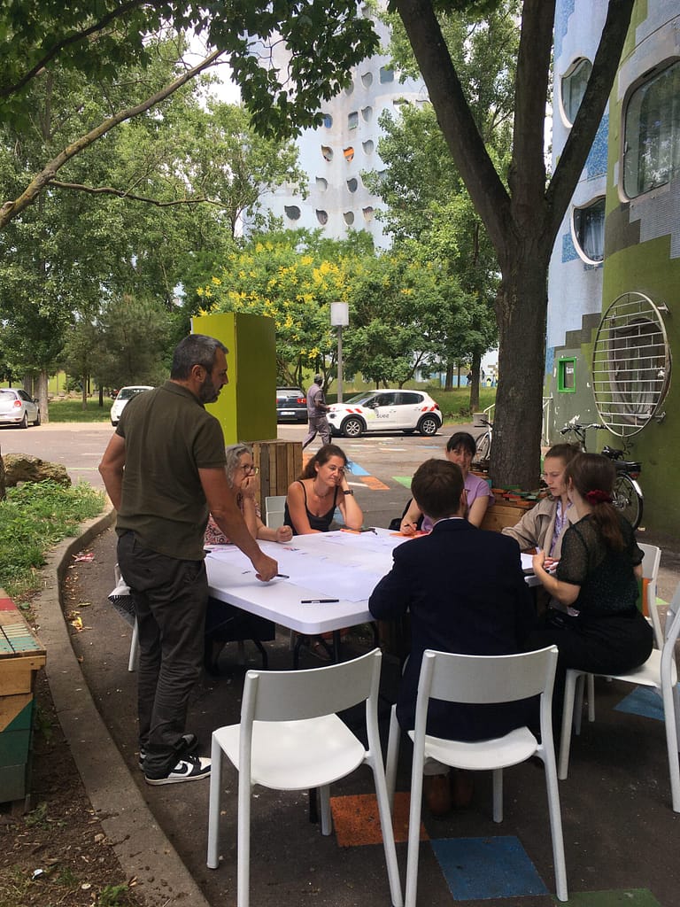 La photo est prise en extérieur, on voir une demi douzaine de personnes assises autour d'une table sous un arbre, semblant réfléchir autour d'un plan à côté d'un bâtiment