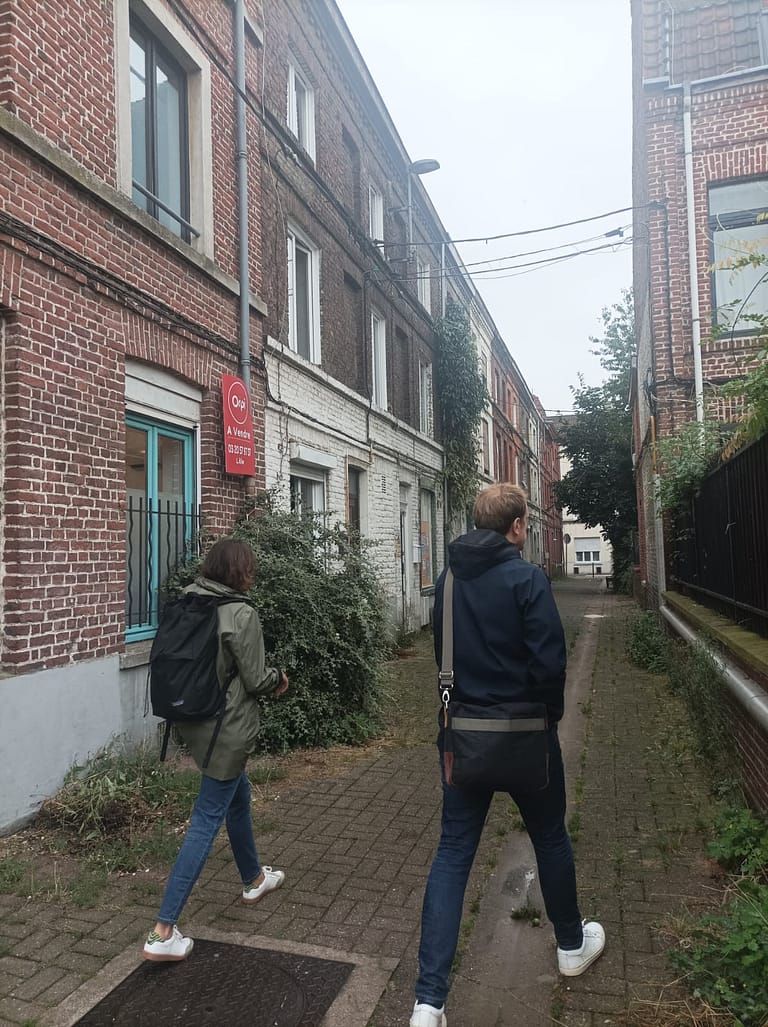 L'image montre 2 personnes marchant dans une rue pavée, encadrée de maisons à 2 étages en brique. Il fait gris