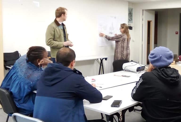 L'image montre 8 personnes assises autour d'une table en intérieur, qui regardent une personne debout au tableau expliquer des cartes colées