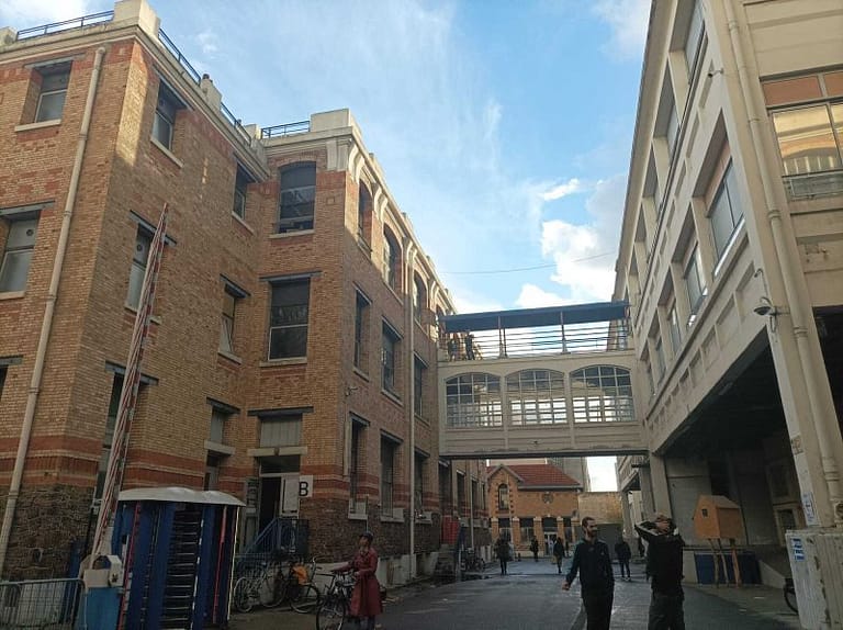 L'image montre les anciennes usines, des bâtiments de 3 étages enbriques avec une passerelle entre les 2. Des personnes à vélo et à pied cheminent en bas