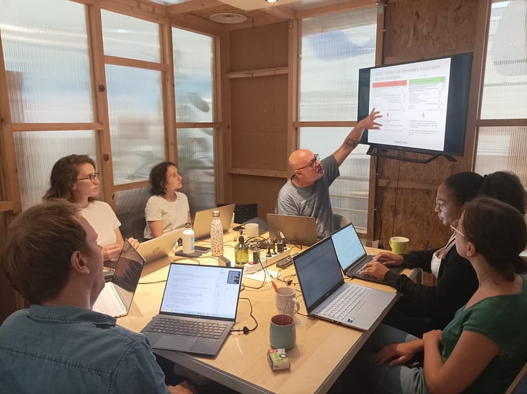 L'image montre 7 personnes assises autour d'une table en intéreur, avec une des personnes qui présentent un powerpoint sur un écran que tout le monde regarde