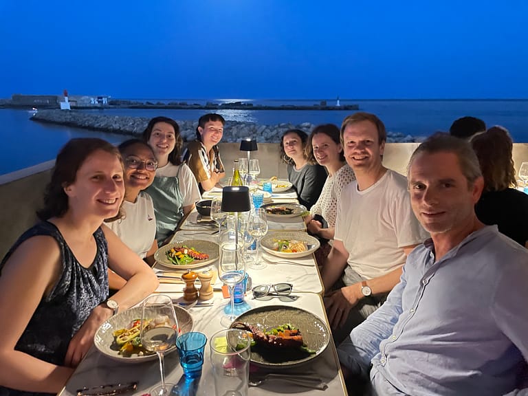 L'image montre 8 personnes souriantes assises autour d'une table dressée, devant la mer