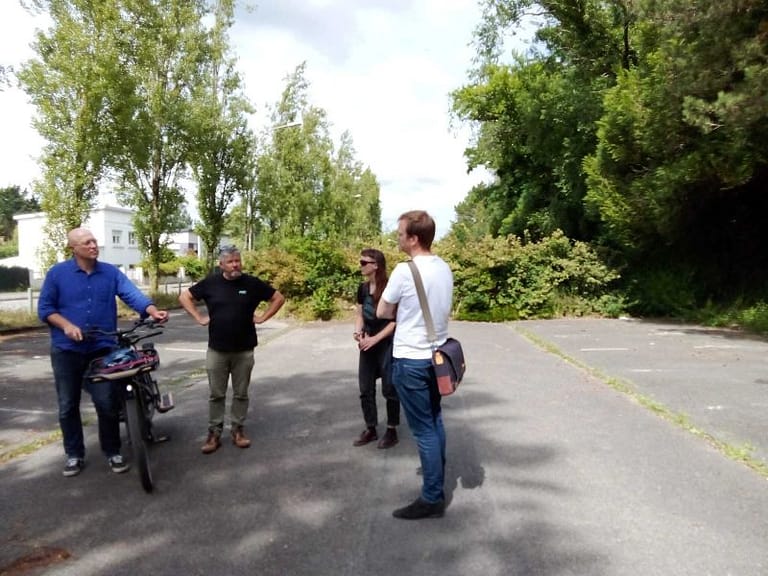 L'image montre 4 personnes debout sur une route au milieu d'immeubles et d'un parc