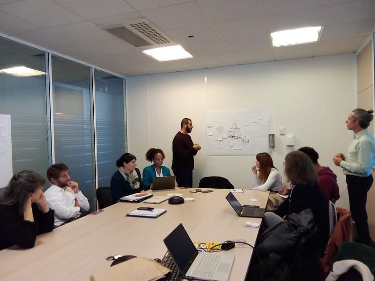 L'image montre 8 personnes assis autour d'une table dans un bureau, écrivant sur leurs ordinateurs. 2 personnes sont edebout au tableau, avec une grande feuille accrocjée au mur