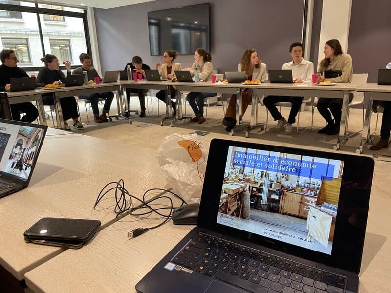 L'image montre une table ronde avec des personnes assises autour devant leurs ordinateurs portables. Un ordinateur au premier plan qui projette un powerpoint avec écrit "immobilier & économie sociale et solidaire"