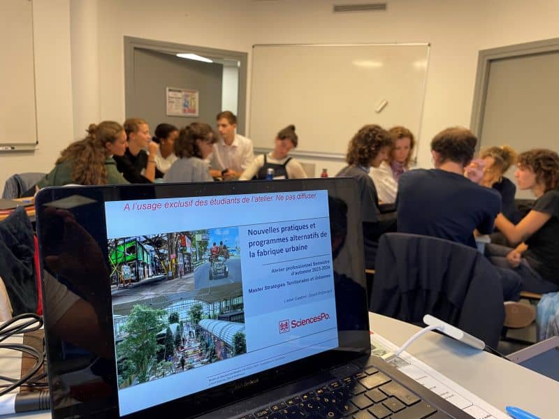 L'image montre une salle de cours avec des étudiants, et au premier plan un ordinateur qui projette un powerpoint "nouvelles pratiques et programmes alternatifs de la fabrique urbaine"