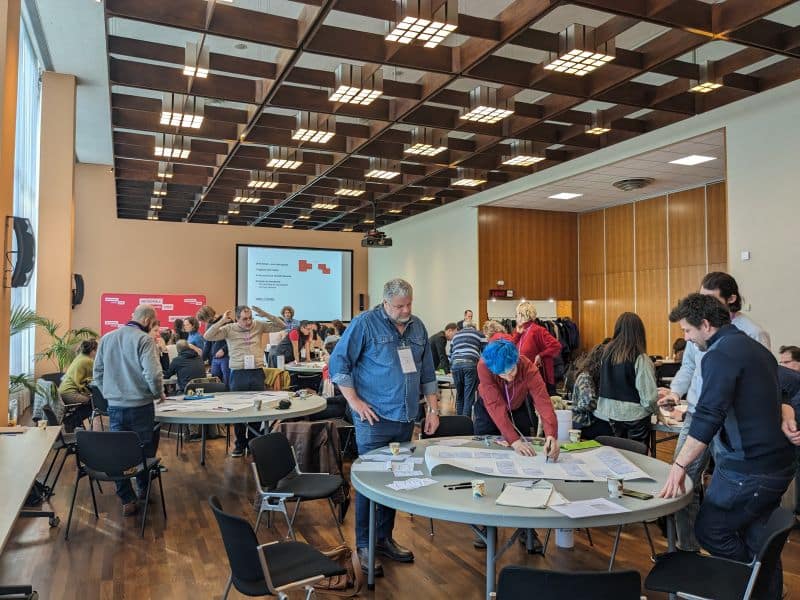 L'image montre une salle au plafond haut avec 5 tables rondses acec des personnes debout autour qui écrivent sur des grandes feuillles de papier.