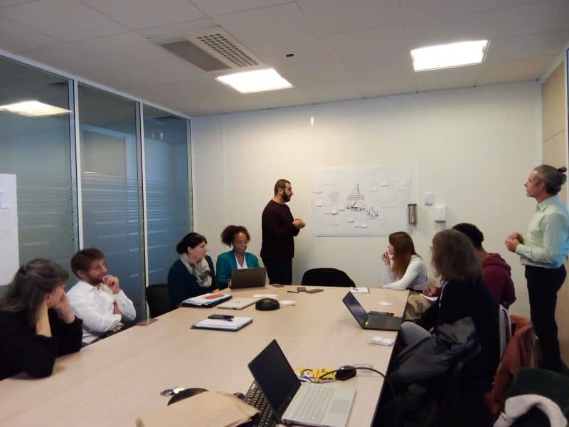 L'image montre 8 personnes assis autour d'une table dans un bureau, écrivant sur leurs ordinateurs. 2 personnes sont edebout au tableau, avec une grande feuille accrocjée au mur