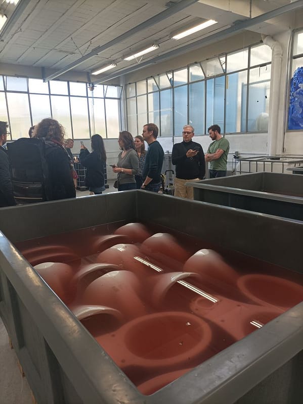 L'image montre au premier plan un bac avec des cuvettes de wc trempée dansun liquide rouge, et au deuxième plan 8 personnes écoutant une 9eme personne parler,d ans un hangar lumineux