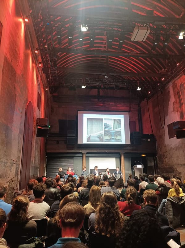L'image montre une conférence avec 6 personnes sur scène devant un parterre de personnes assises, au sein d'une ancienne église à haut toit en coque de bateau inversée