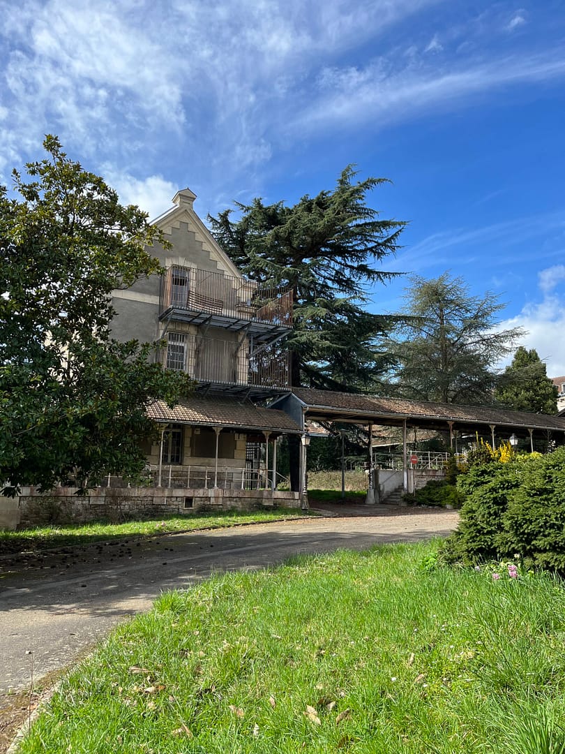 L'image montre au premier plan du gazon, puis un chemin, et enfin une haute maison entourée d'arbres haut avec un ciel bleu
