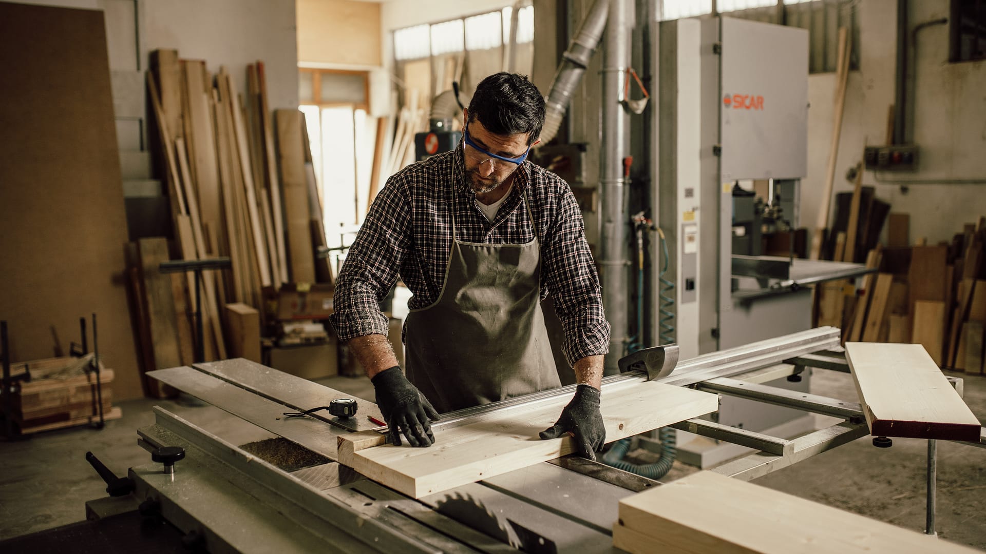 L'image montre une personne en chemise à carreaux et tablier découper du bois dans un atelier, avec des lunettes de protections et des gants, à l'aide d'une scie sauteuse